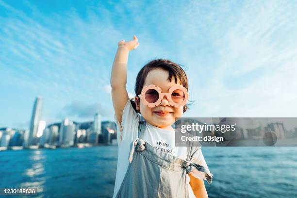 happy little asian girl with flower-shaped sunglasses smiling joyfully while having fun outdoors and imagines flying in the air against a clear blue sky and urban city skyline - hong kong girl stock-fotos und bilder