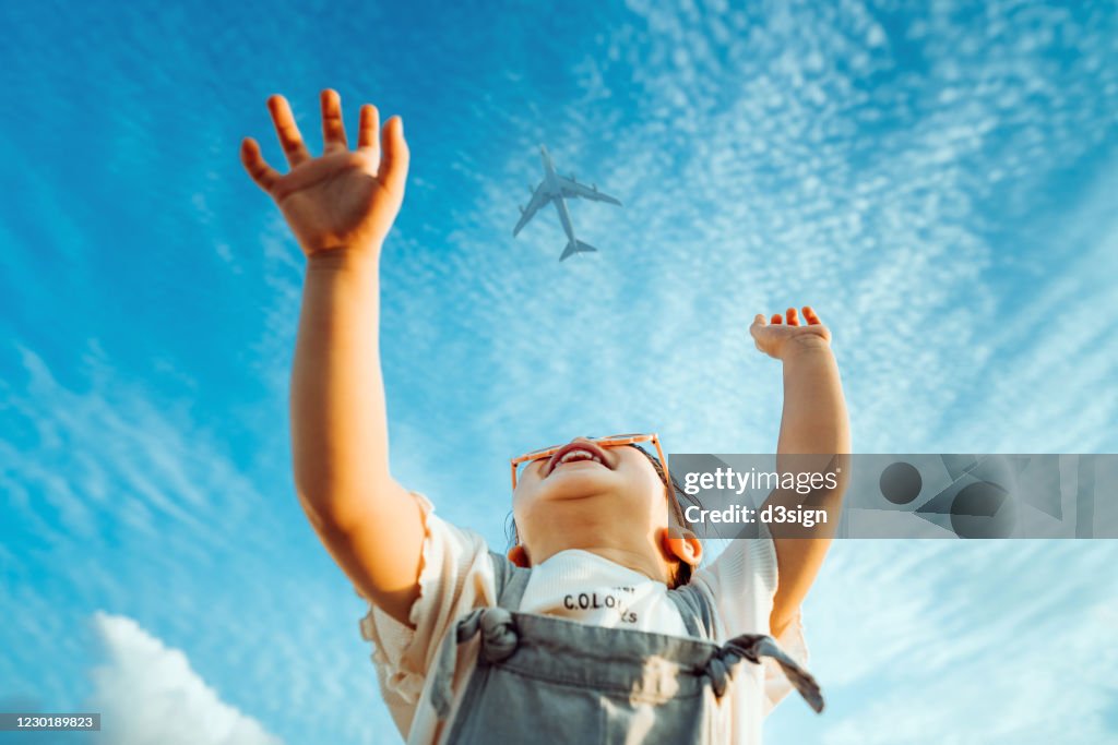 Happy little Asian girl with flower-shaped sunglasses smiling joyfully and raised her hands waving to the aeroplane in the clear blue sky