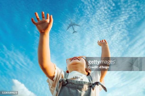 happy little asian girl with flower-shaped sunglasses smiling joyfully and raised her hands waving to the aeroplane in the clear blue sky - neugierde stock-fotos und bilder