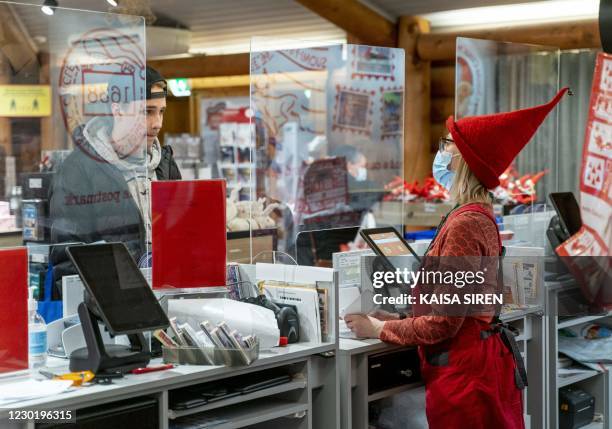 An employee wearing a Christmas hat and face mask to protect against the coronavirus serves a customer through a plexiglass screen at Santa Claus'...