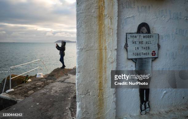 Woman takes picture of the sea next to a shelter with an artworks that reads ' Don't Worry We're All In This Together' seen at the Vico bathing...