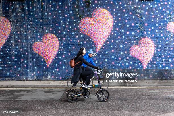 Couple is seen riding bicycles on Sunday sunny afternoon in Southbank Borough area in front of hearts graffiti as the UK's government introduced...