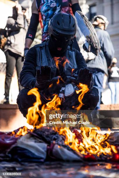 Member of an anti-fascist group warms his hands next to a burning American flag on the steps of the Colorado State Capitol on January 20, 2021 in...