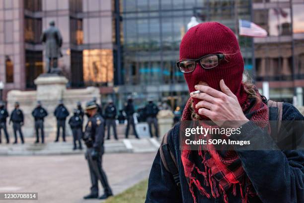 Demonstrator wearing a balaclava smokes a cigarette in front of the Ohio Statehouse with a line of Ohio State Troopers behind him. Activist groups in...