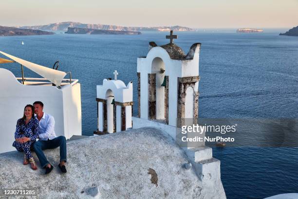 Honeymooning couple sit atop a church overlooking the village of Oia on Santorini Island, Greece. Santorini Island is without a doubt one of the most...