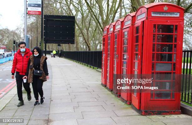 Couple wearing facemasks walk past 4 red telephone boxes alongside Hyde Park. England remains under lockdown as the Prime Minster Boris Johnson...