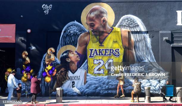 People gather in front of a mural of former Los Angeles Laker Kobe Bryant and his daughter Gianna, both with a set of wings, by artist sloe_motions...