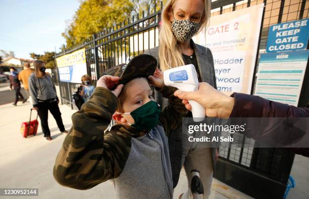 First grade student Baron White gets his hat into position to have his temperature taken by teachers aide Firoozeh Borjian as he arrives with his Mom...