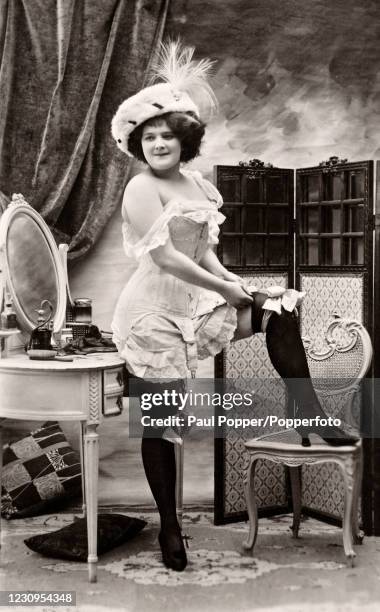 Provocative vintage French postcard featuring a young woman undressing in her boudoir wearing a large plumed hat and black stockings, published in...
