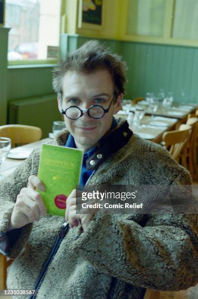 Ralph Fiennes sitting in a cafe, wearing Harry Potter glasses and reading "Quidditch through the Ages" that was sold in support of Red Nose Day 2001.