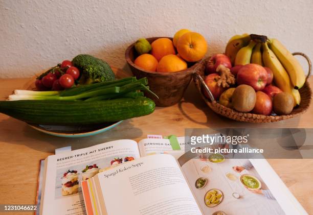February 2021, Berlin: Different kinds of vegetables and fruit lie in baskets on a table in an apartment in Prenzlauer Berg. Two cookbooks lie open...