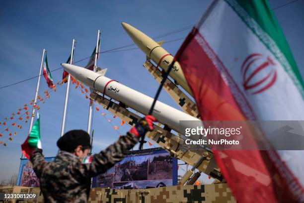 An Iranian young boy wearing an Islamic Revolutionary Guard Corps uniform holds an Iran flag while Iran-made, Dezful medium range ballistic missile...