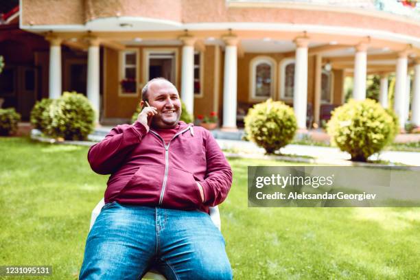 happy male using phone while relaxing on backyard grass park - fat man sitting stock pictures, royalty-free photos & images