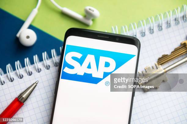 In this photo illustration, a SAP logo seen displayed on a smartphone with a pen, key, book and headsets in the background.