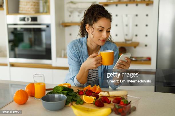 jonge vrouw die haar mobiele telefoon gebruikt terwijl het voorbereiden van fruitmoothie in de keuken - mengen stockfoto's en -beelden