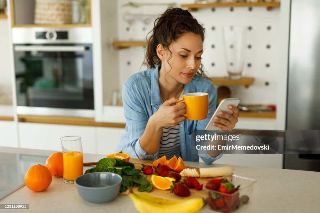 Jovem usando seu celular enquanto preparava smoothie de frutas na cozinha