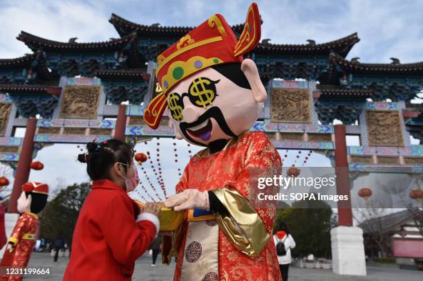 Kid touching the ornament "jinyuanbao" held by a staff wearing clothes of "God of wealth" to attract tourists in Fuyang ecological paradise square....