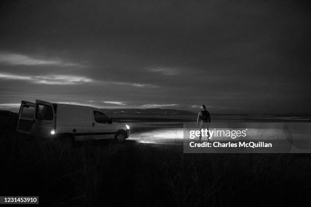 Surfer Al Mennie is illuminated by his vans headlights as the light falls on Downhill Strand before he takes to the water on March 1, 2021 in...