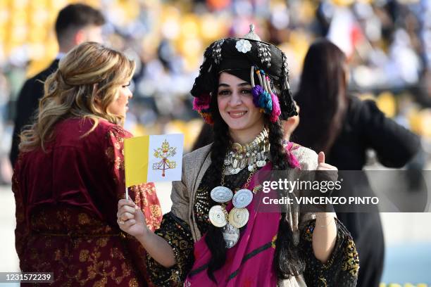 Young woman dressed in traditional Assyrian clothing holds a flag of the Holy See and gestures as she waits for the arrival of Pope Francis at the...