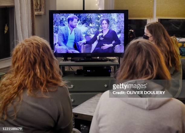 This photo illustration shows people wearing face masks, watch a televised conversation between Britain's Prince Harry with his wife Meghan Markle...