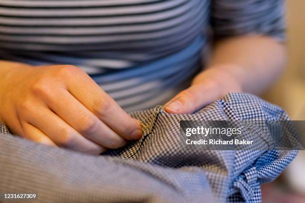 Young dressmaker woman uses a needle and thread to sew together a home-made dress that she's created from a pattern in her home, on 6th March 2021,...