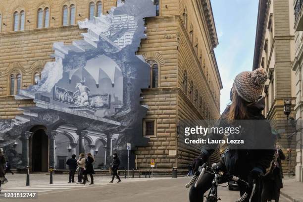 Woman on a bicycle looks up at the facade of Palazzo Strozzi with the installation by artist JR The Wound at Palazzo Strozzi on March 19, 2021 in...