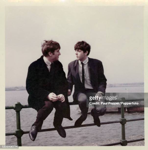 Vintage snapshot featuring John Lennon and Paul McCartney of the Beatles sitting on a railing beside the River Mersey during filming for the BBC TV...