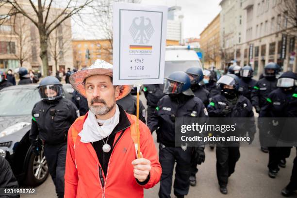 March 2021, Berlin: A man wearing an aluhut and carrying a Basic Law sign walks in front of police officers during a demonstration against the Corona...