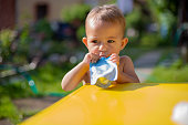 cute little caucasian baby eating fruit puree in pouch and looking into the camera in front of the yellow table. close up, on the background is  a green garden on a sunny day in blur