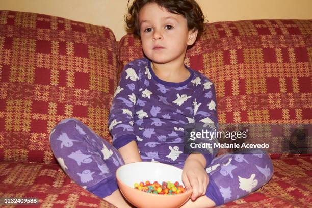 toddler boy sitting on a couch eating brightly covered cereal out of a big bowl he is holding. he is in his pajamas which are purple and white. - boy pajamas cereal stock pictures, royalty-free photos & images