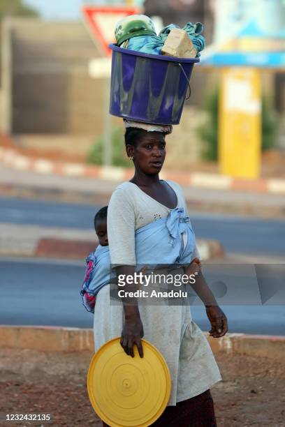 Woman vendor carries a baby and goods in a bucket over her head in Bamako, Mali on April 20, 2021. Malian women, struggling for water, food and...