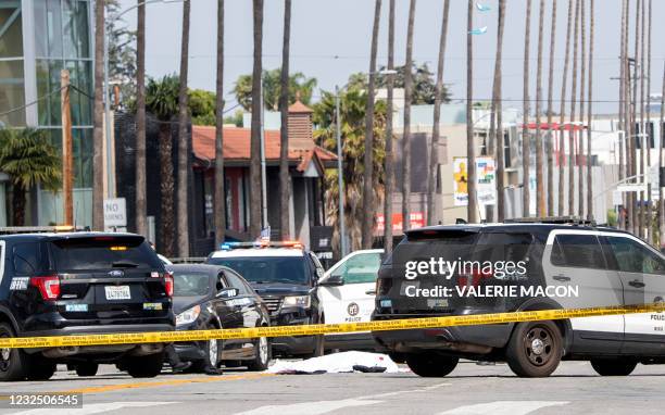 Body covered in a white sheet lies next to a black vehicle covered with stickers and a shattered driver side window, as police cars block traffic at...