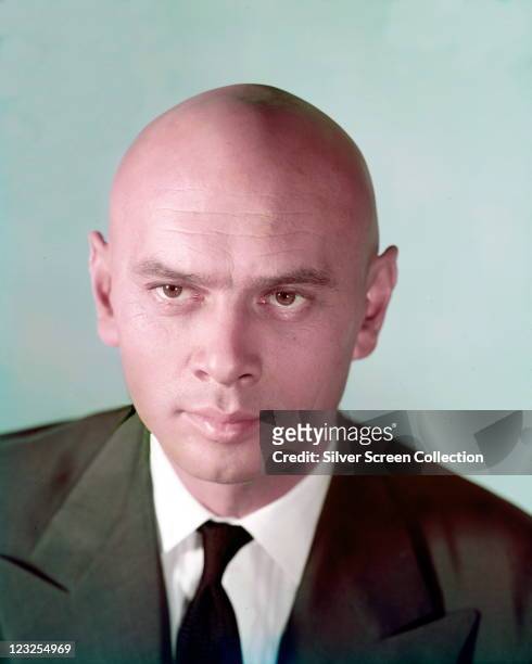 Yul Brynner , Russian-born US actor, wearing a green jacket with a white shirt and a black tie in a studio portrait, against a light blue background,...