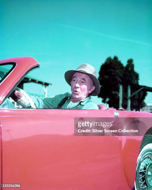 Walter Brennan , US actor, wearing a hat while driving a red sports car, with trees and blue sky in the background, circa 1965.