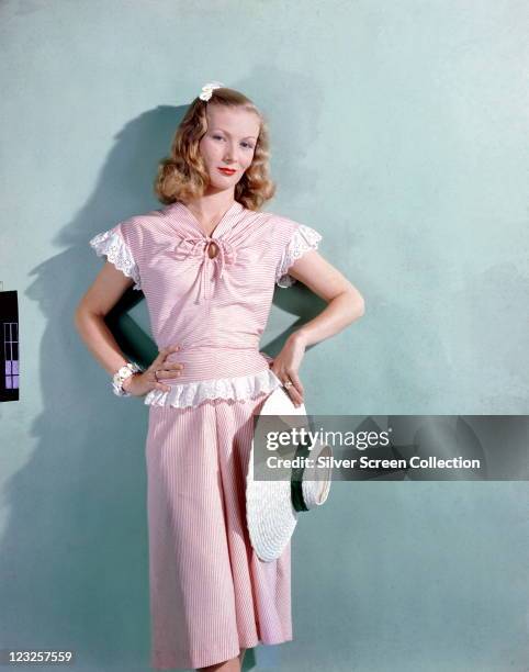 Veronica Lake , US actress, wearing a pink gingham dress with white lace trim, holding a white wide-brimmed straw hat with a green band, in a studio...