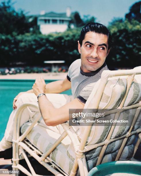 Tyrone Power , US actor, wearing a white-and-blue striped t-shirt as he turns to look over his shoulder as he sits on a bamboo pool chair beside a...
