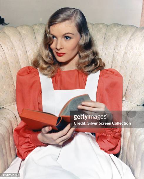 Veronica Lake , US actress, wearing a red blouse beneath a white apron dress, sitting in an armchair as she reads a book, circa 1955.