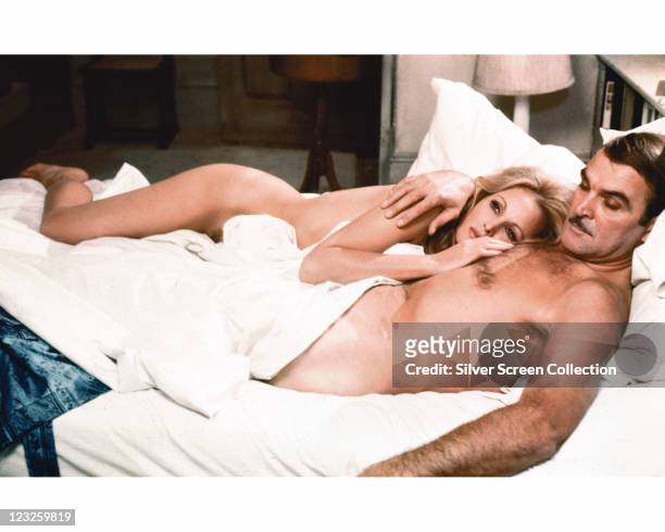 Ursula Andress, Swiss actress, laying naked on a bed with Stanley Baker , British actor, who is covered from the waist down by a white sheet in a...