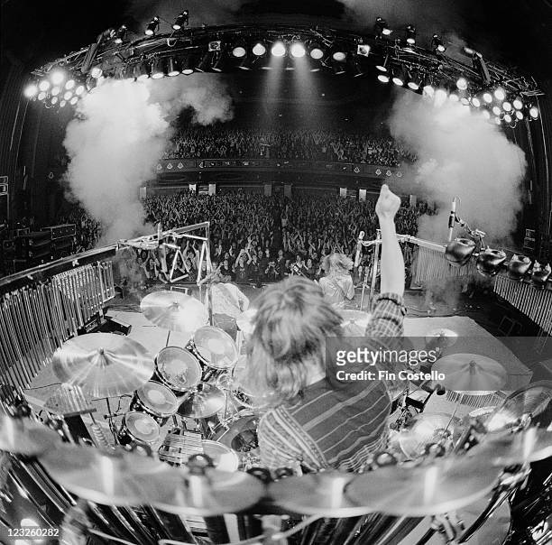 View looking from the back of the stage, looking out over drummer Neil Peart sitting at his drumkit with an arm raised aloft, guitarist Alex Lifeson...