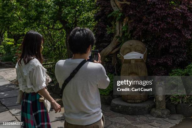 Couple wearing face masks pause to photograph a deity adorned with a face mask in the grounds of a Shinto shrine on May 4, 2021 in Kamakura, Japan....