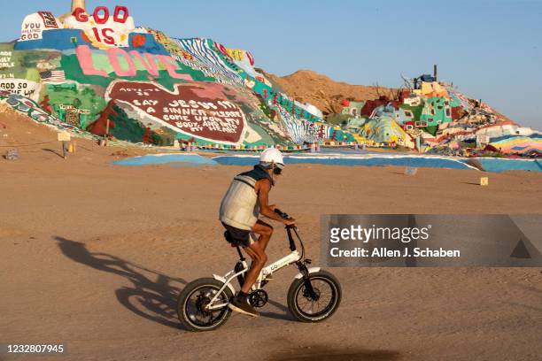 Slab City, CA A person rides their bike as tourists visit Salvation Mountain, hillside Christian monument paying homage to Jesus Christ and was...