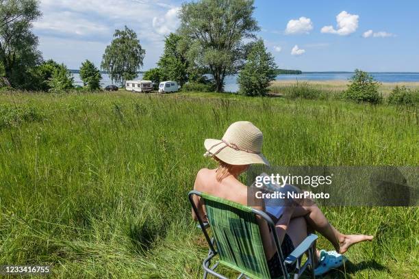 Woman reading a book in a nature in front of her self converted campervan wearing a straw hat is seen at Szeroki Ostrow peninsula, Sniardwy lake,...