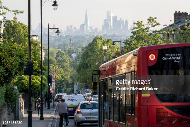 With the London skyline in the far distance, a bus and morning traffic descend Gypsy Hill in Crystal Palace, on 16th June 2021, in London, England.