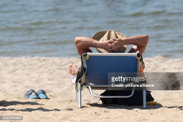 June 2021, Saxony, Boxberg: A man with a sun hat is lying on a sun lounger on the shore of the Bärwalder See on the beach. Photo: Sebastian...