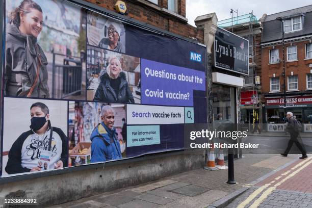 South Londoner wearing a face covering walks past a governement NHS billboard giving a link for official information relating to Covid vaccine facts,...
