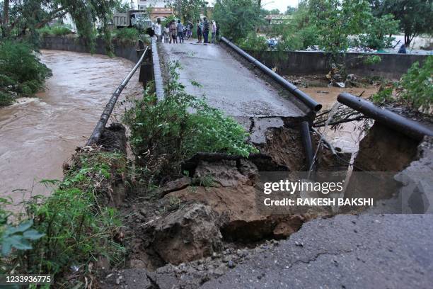 Local residents stand on the edge of a road bridge that was damaged by flash floods after heavy monsoon rains on the outskirts of Jammu on July 12,...