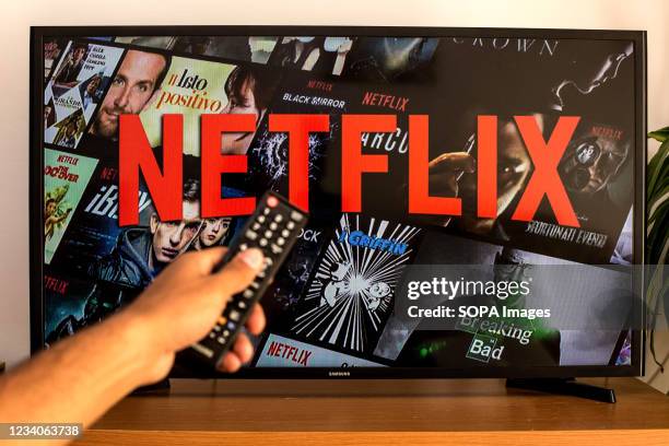 In this photo illustration a close-up of a hand holding a TV remote control seen displayed in front of the Netflix logo on a tv in the background.