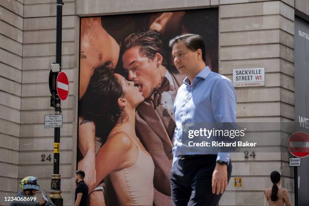 Londoners pass beneath a tall advertising billboard, by menswear retailer Suitsupply, of an intimate kissing couple on Covid 'Freedom Day'. This date...