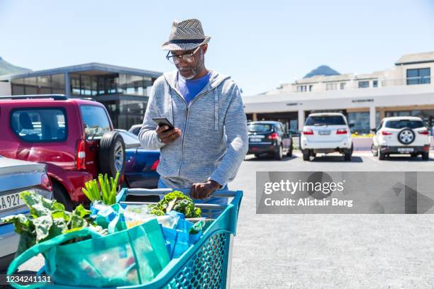 mature african man pushing shopping cart while looking at smart phone - happy consumer on phone stock pictures, royalty-free photos & images