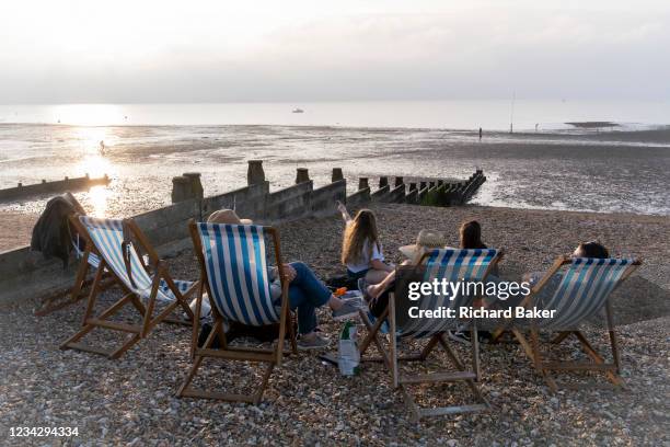 Staycationers sit in deck chairs on beach shingle, to enjoy the calm of a low-tide evening sunset, on 25th July 2021, in Whitstable, Kent, England.